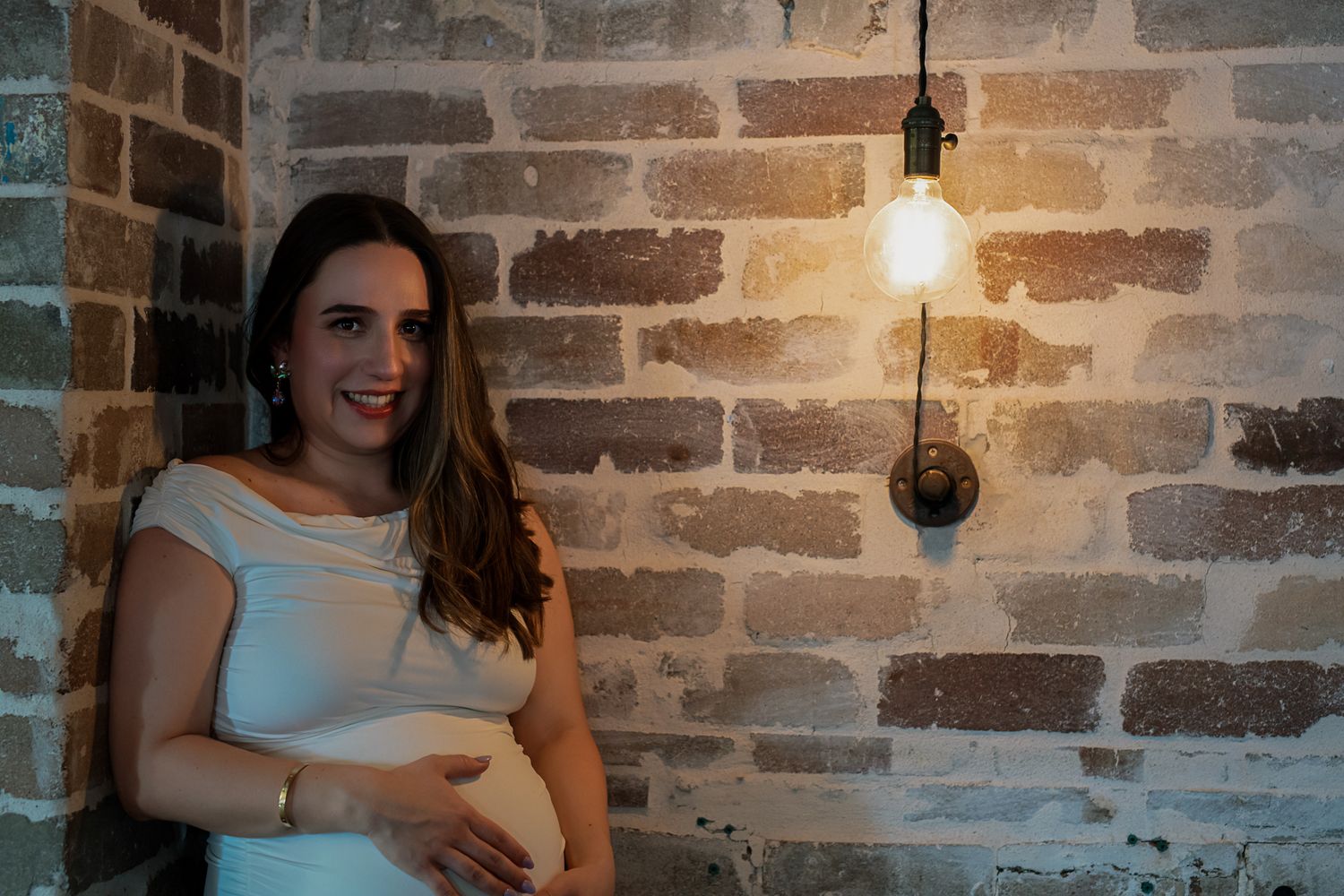 Expectant mother smiling against brick wall with warm lighting from hanging bulb.
