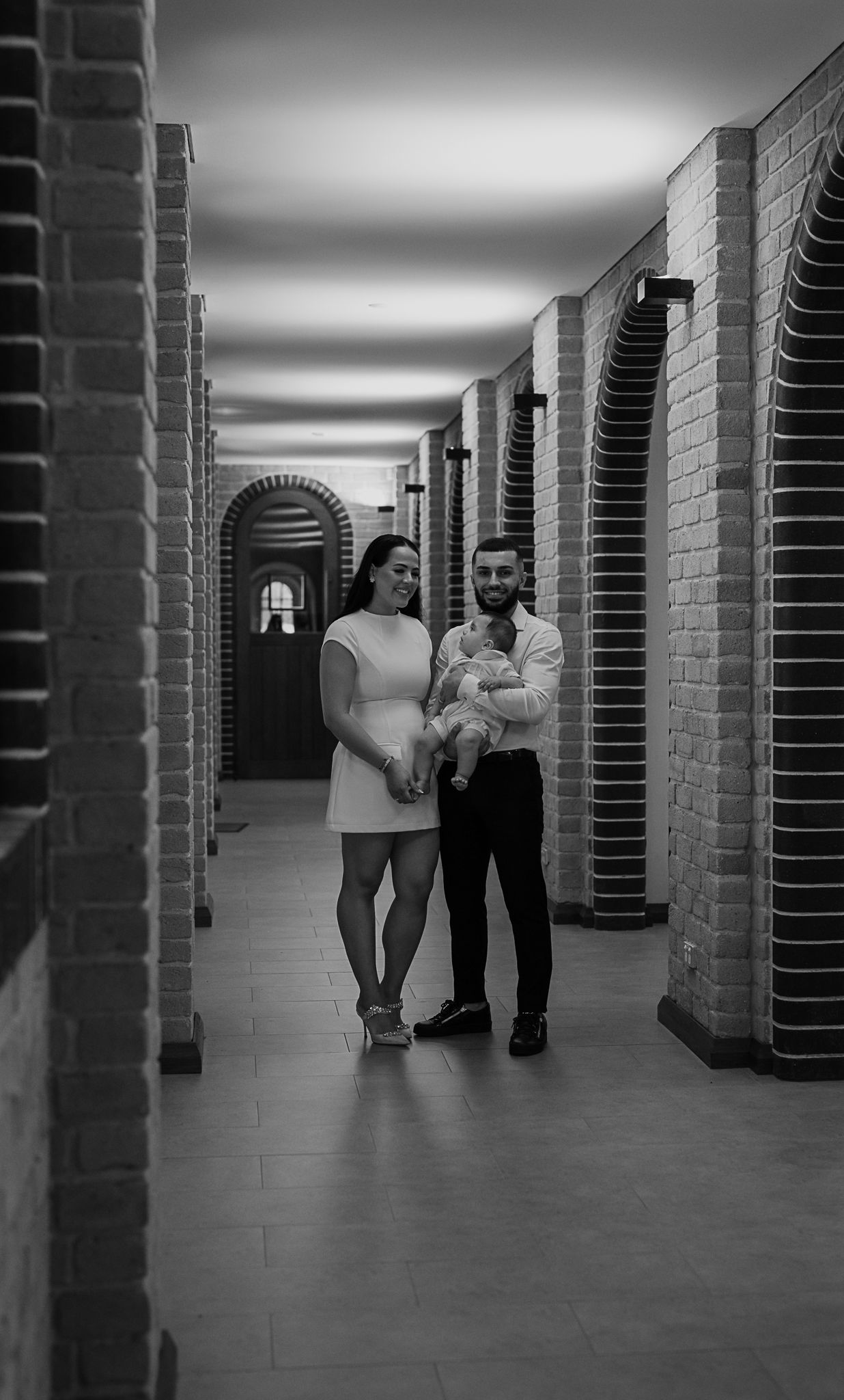 Black and white photo of a couple walking down an arched hallway while holding a baby.