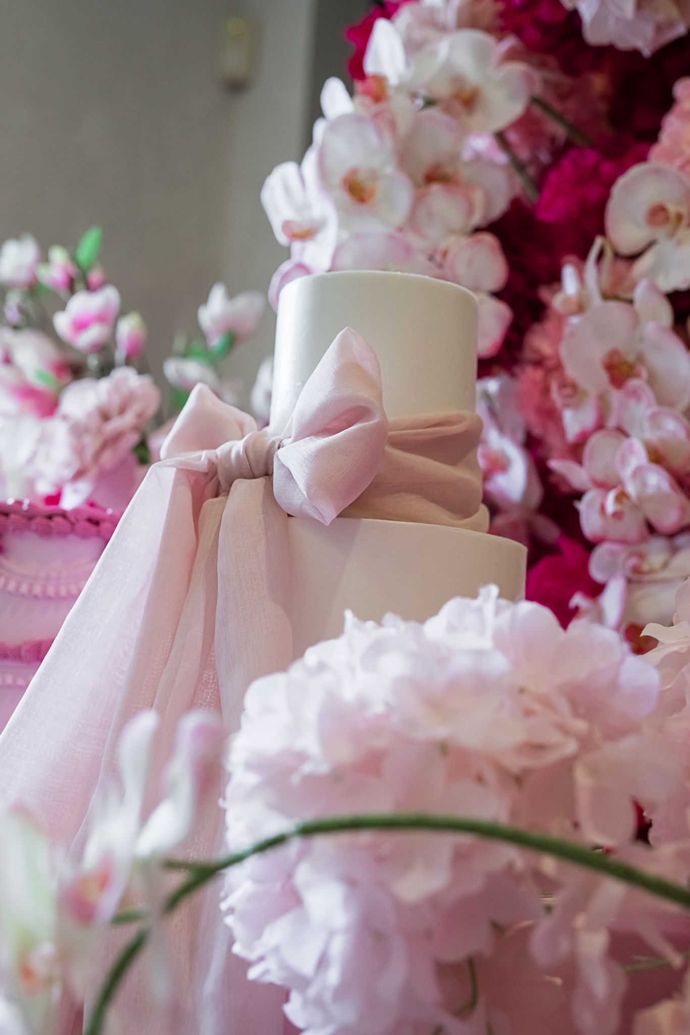 White tiered cake adorned with a pink bow and surrounded by delicate pink flowers.
