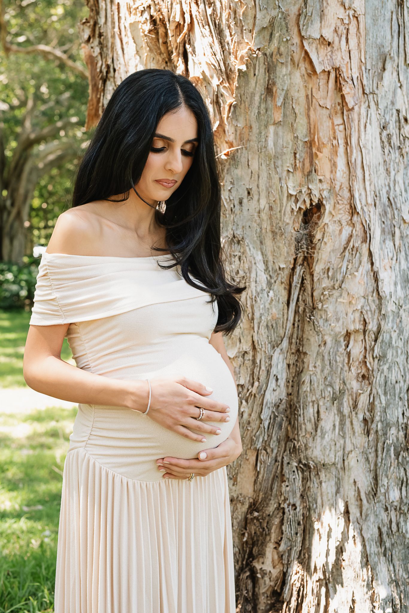 Expectant mother in off-shoulder cream dress poses near a tree trunk in a garden setting.