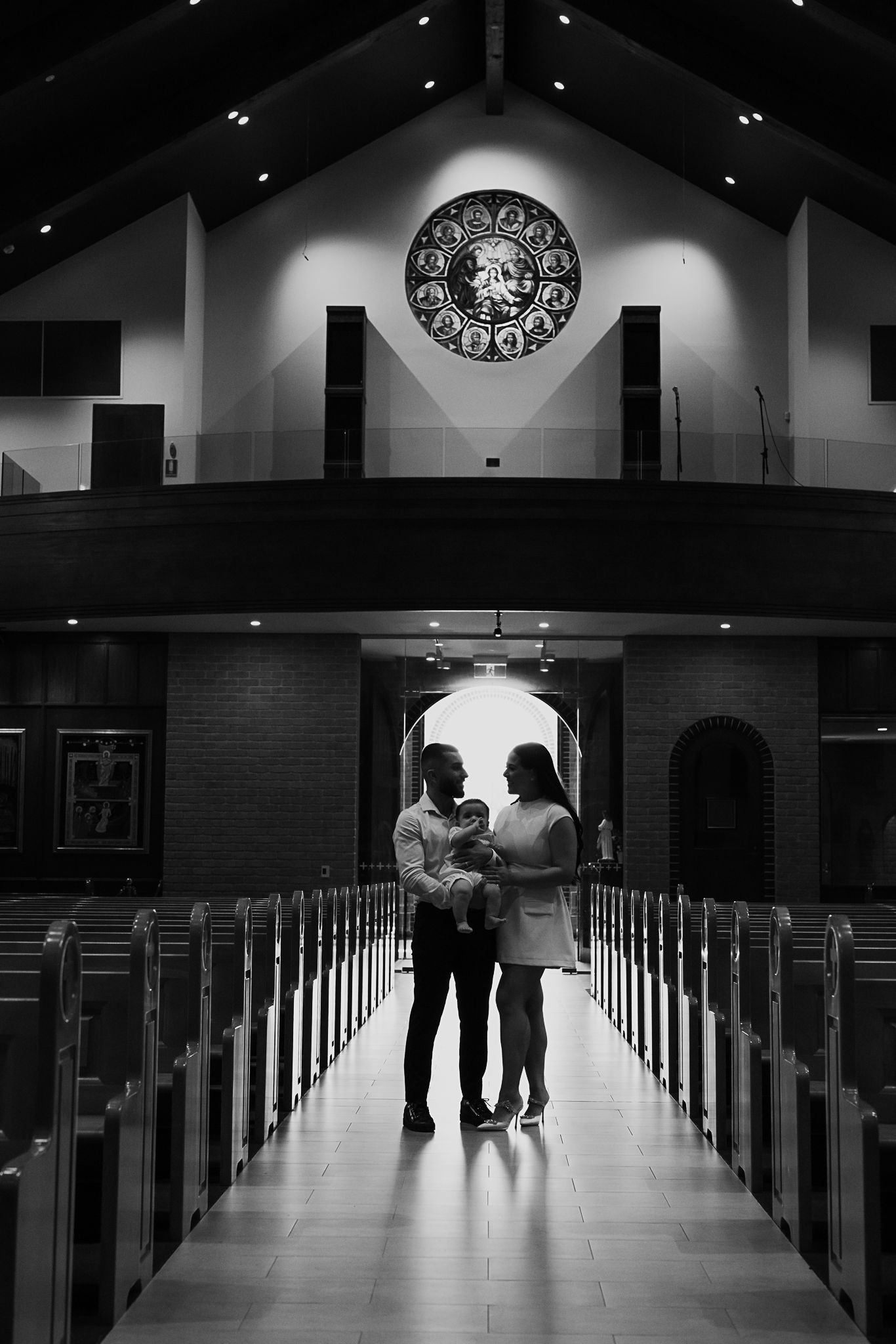 A silhouetted couple with baby stand in a church aisle beneath a decorative circular emblem.