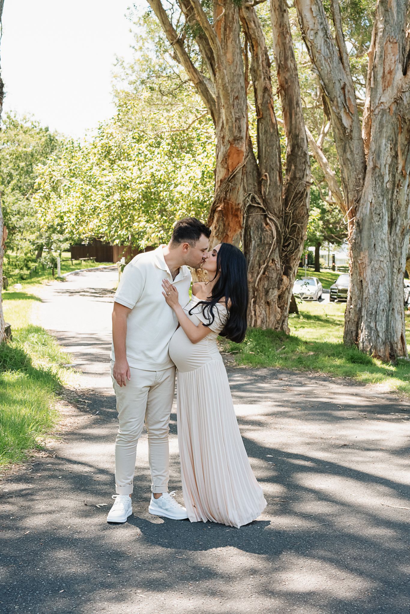 Romantic maternity moment captured between couple in matching white outfits under tall trees.