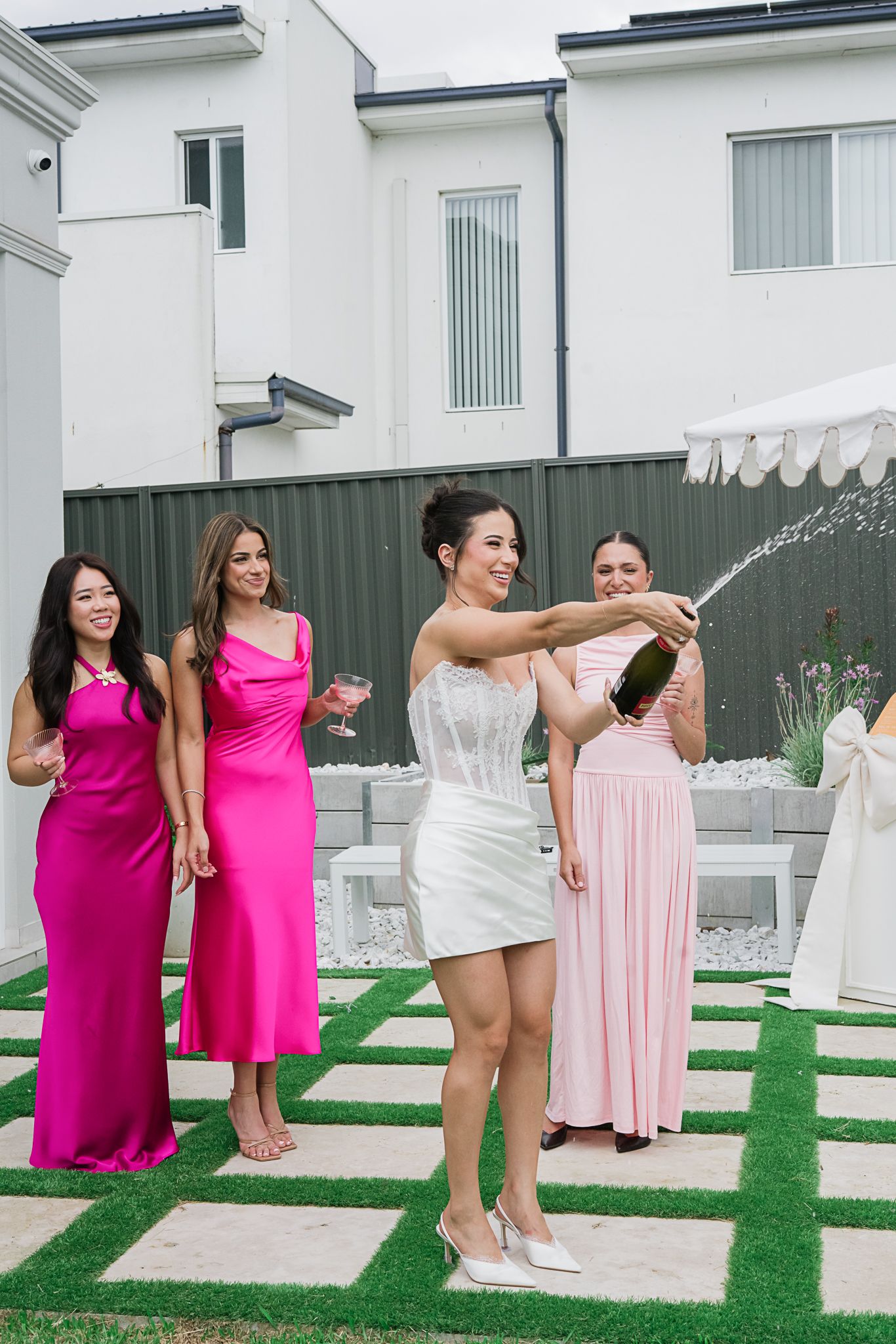Festive champagne spray moment with bridal party in pink and white dresses on garden patio.