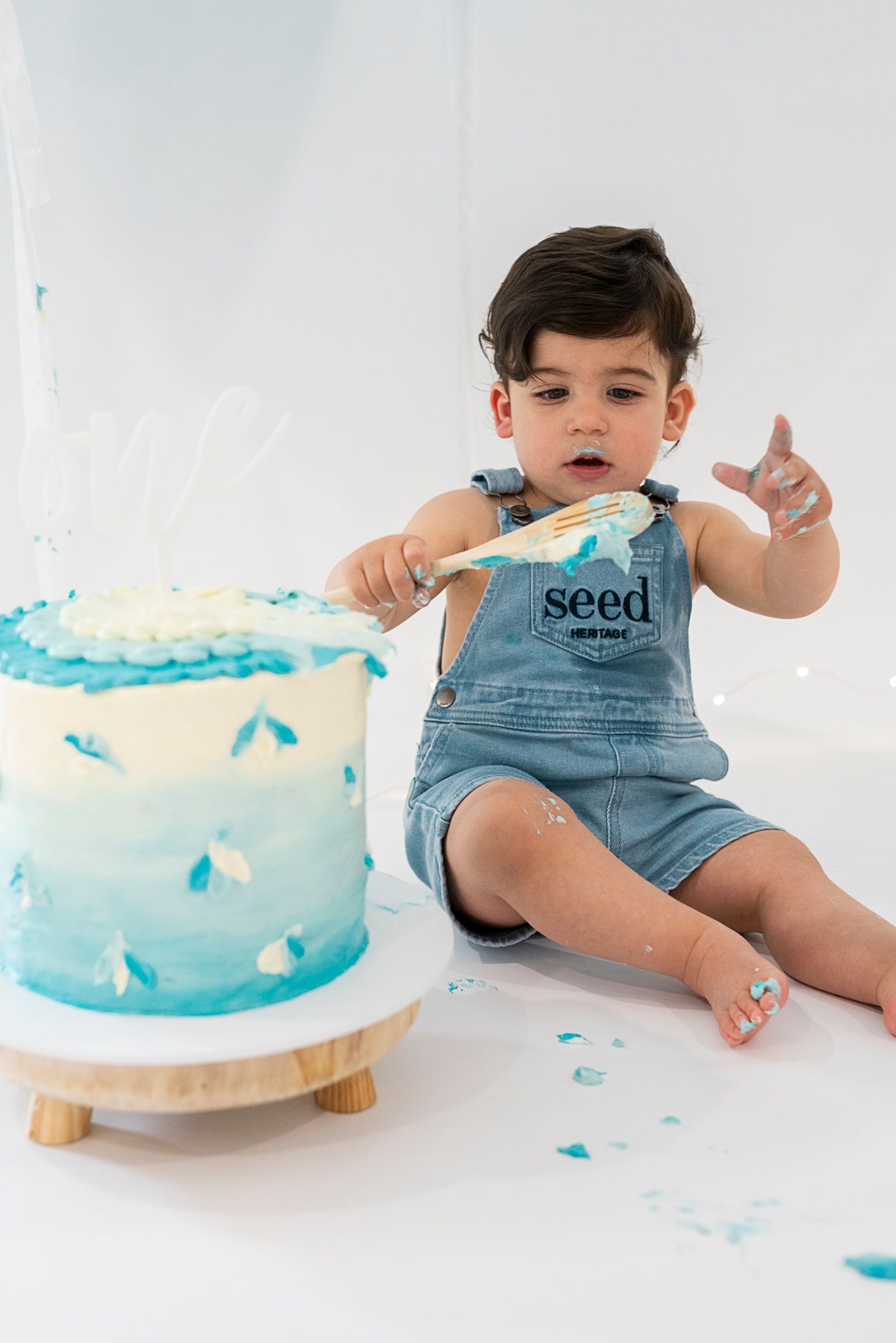 Baby in denim overalls reaches for blue ombre birthday cake during cake smash photoshoot.