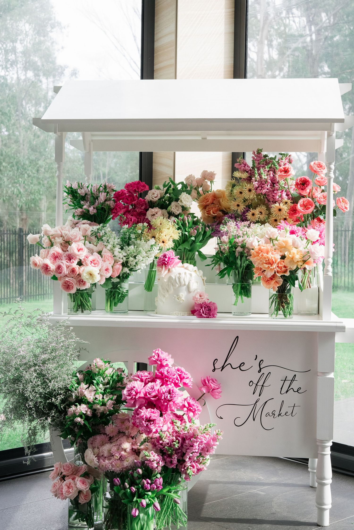 A white market flower stand displaying beautiful bouquets of pink and peach roses with cursive text on the front.