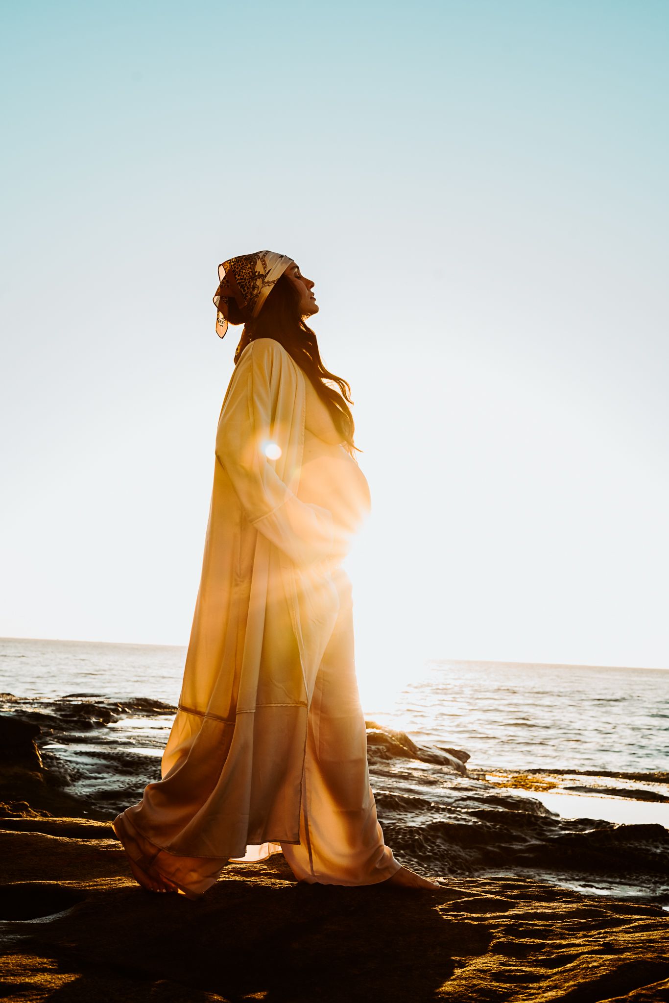 Person in flowing yellow dress standing on coastal rocks at sunset.