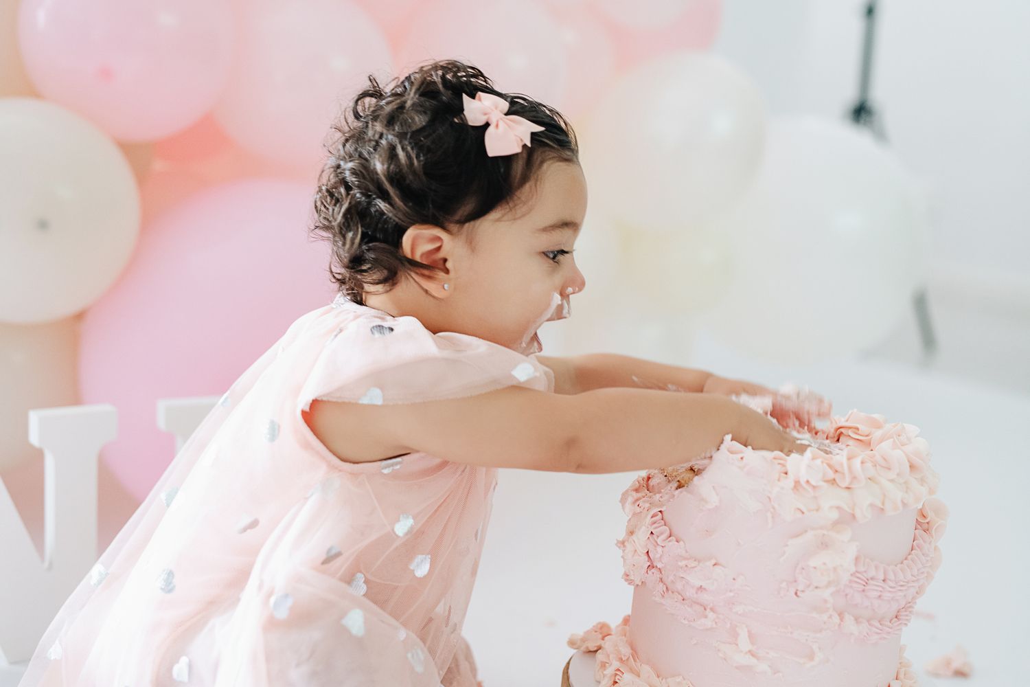 A child in pink dress reaches toward cake during a festive photo session with balloons.