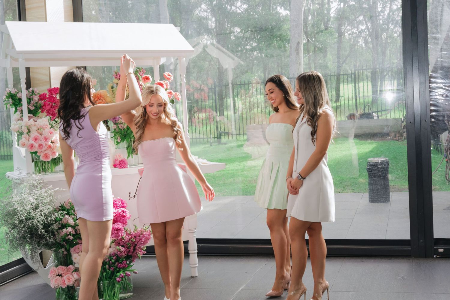 Group of friends in white and pink dresses celebrate joyfully in a sunlit room decorated with pink flowers.