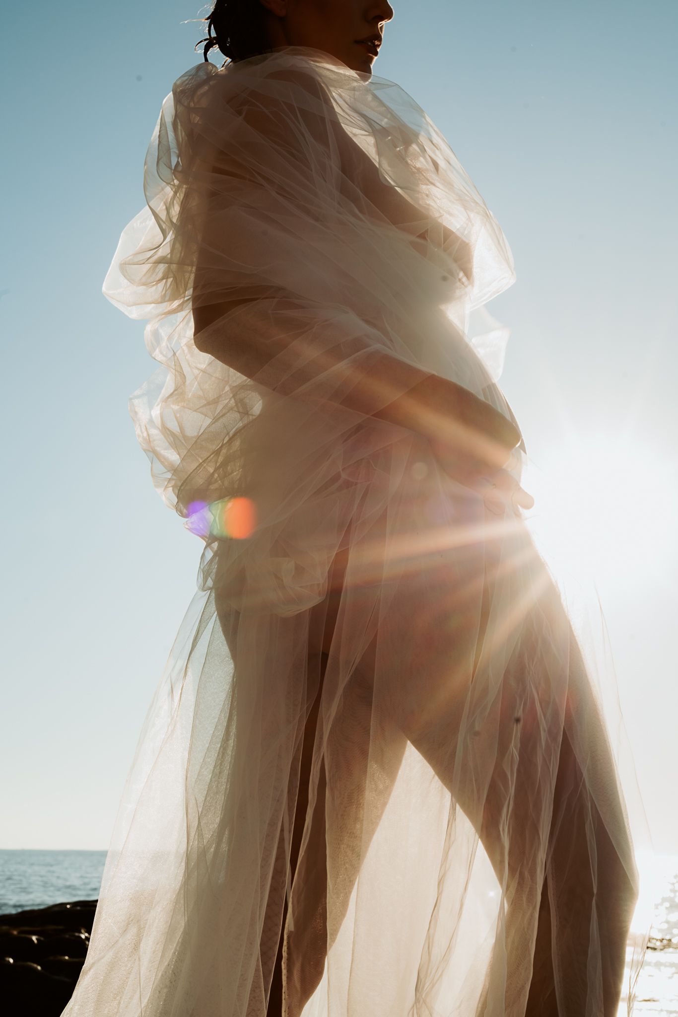 Artistic maternity photograph with sunlight creating lens flares through sheer white fabric.