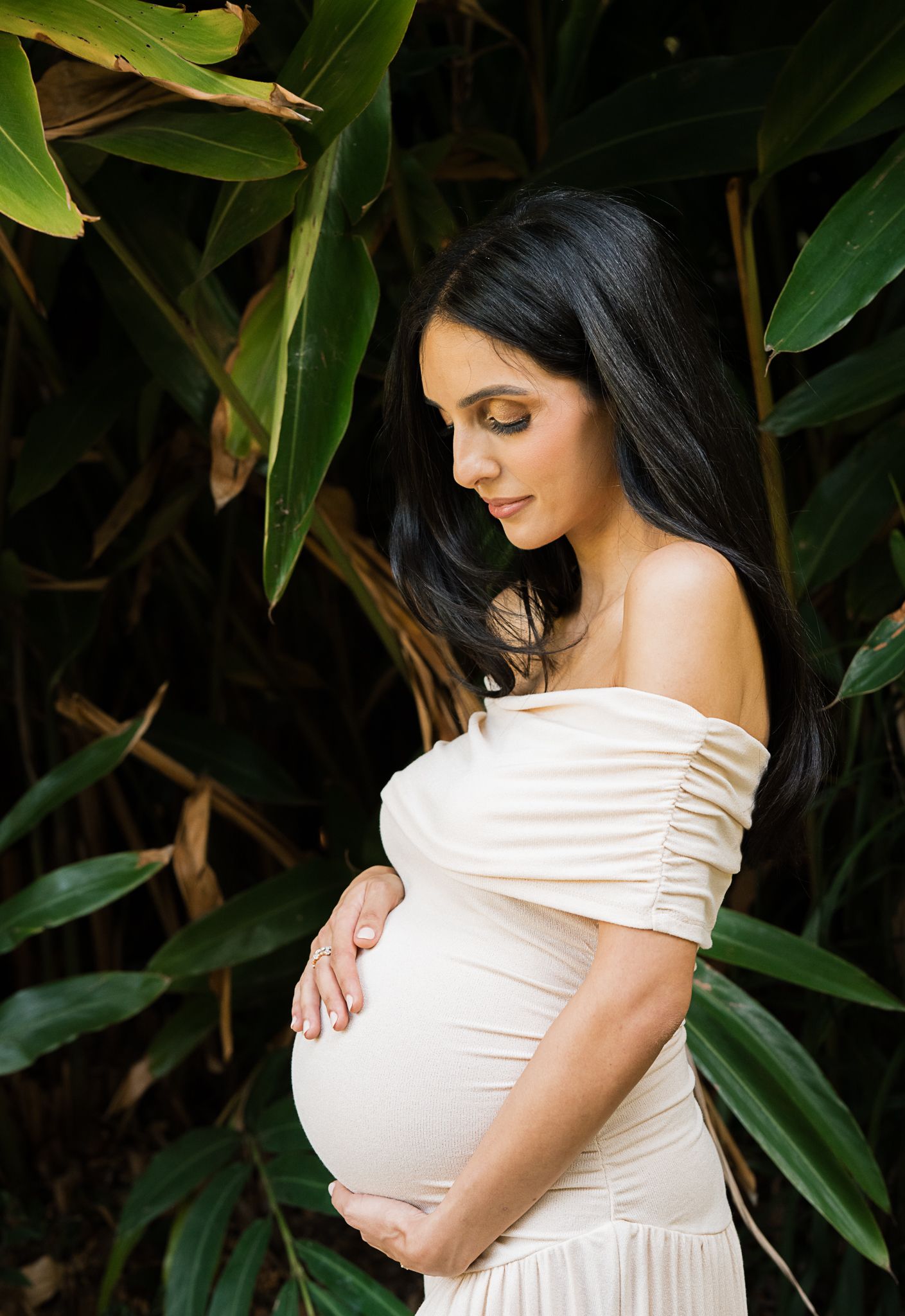 An expectant mother in a white off-shoulder dress stands against lush green foliage in a maternity photo.