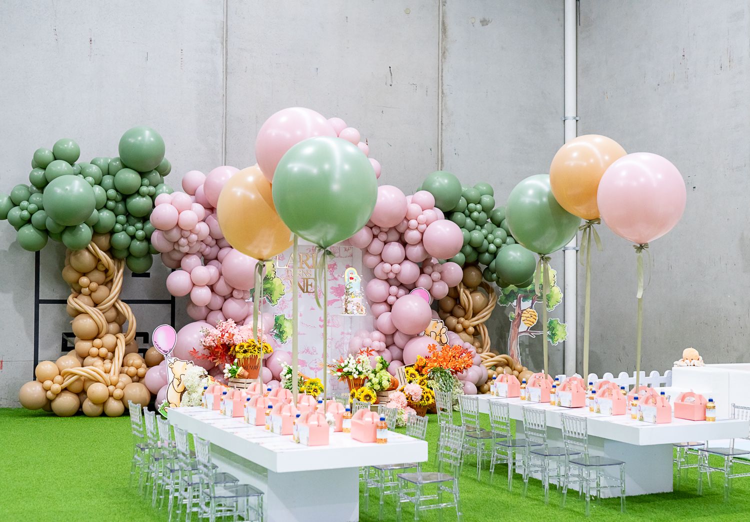 Elegant party setup with pink balloon garland, white tables, and floral arrangements on artificial grass.