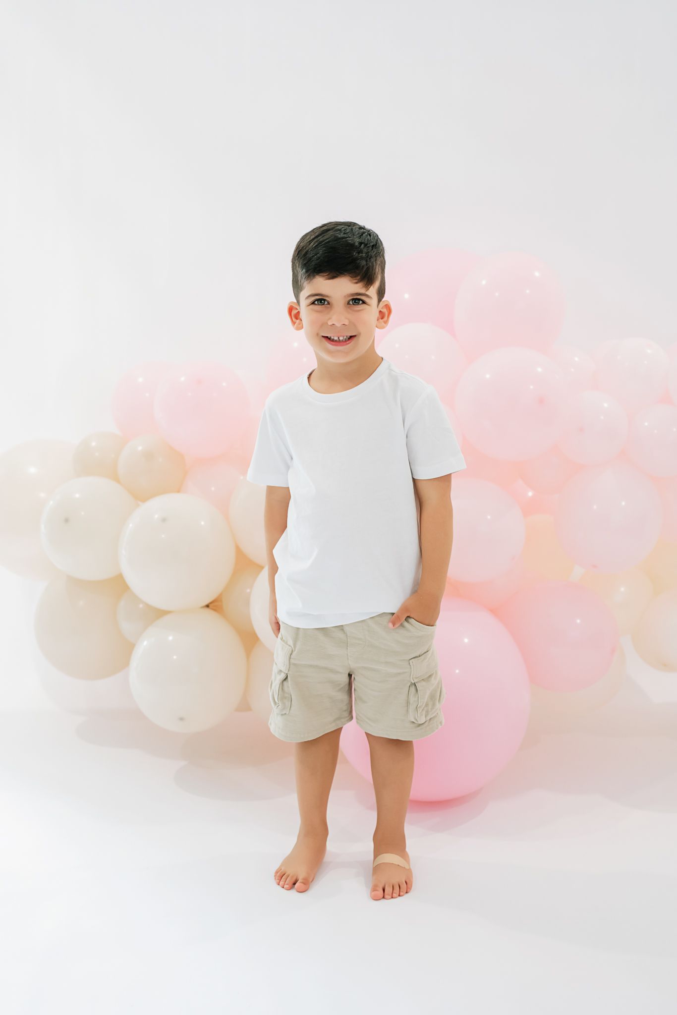 A boy in white shirt and khaki shorts stands barefoot against pink balloon background.