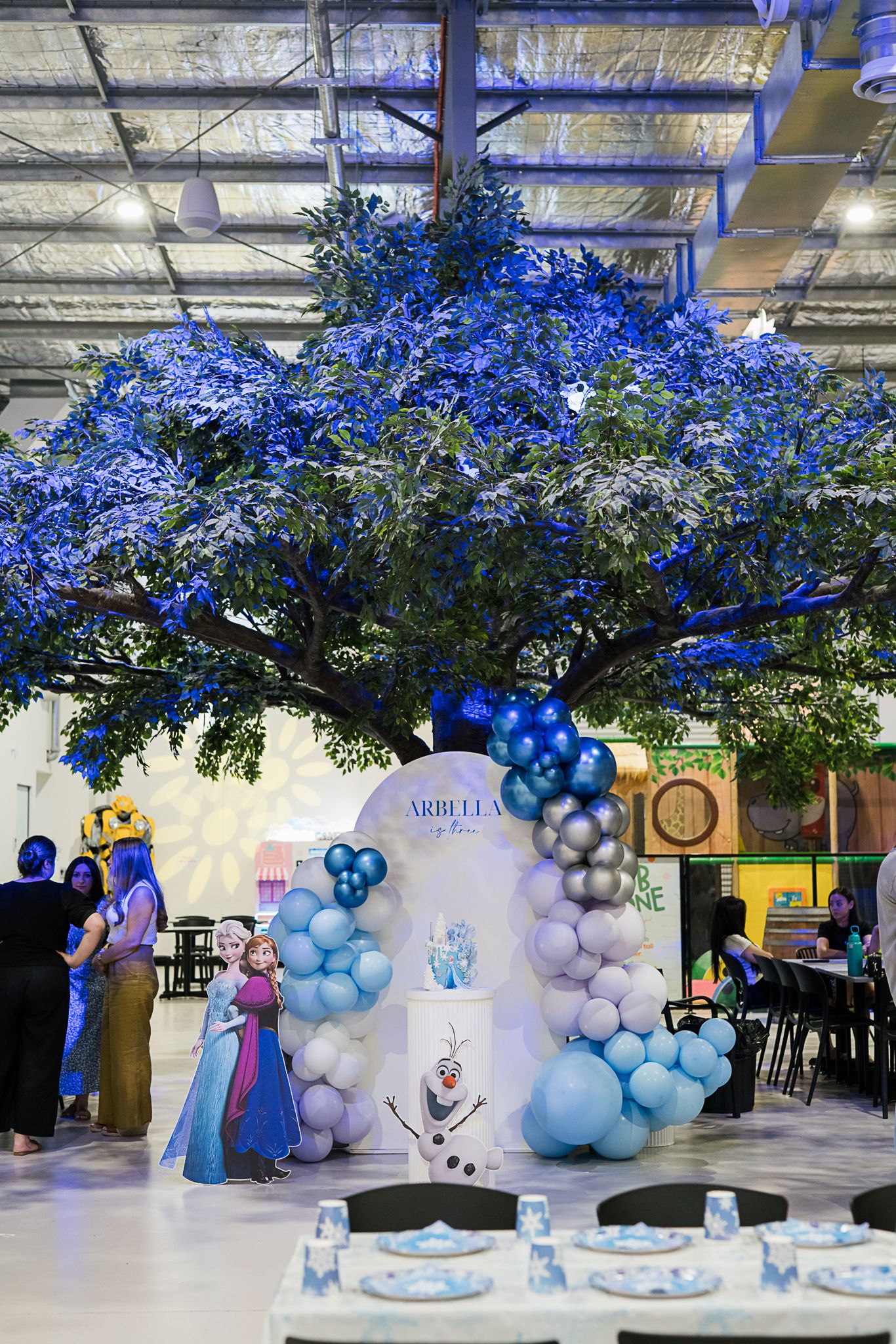 Indoor party venue with illuminated blue tree, balloon decorations, and themed party setup.