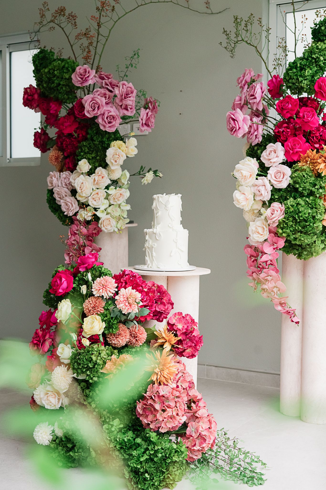 White wedding cake surrounded by lush pink and green floral arrangements on white pedestals.