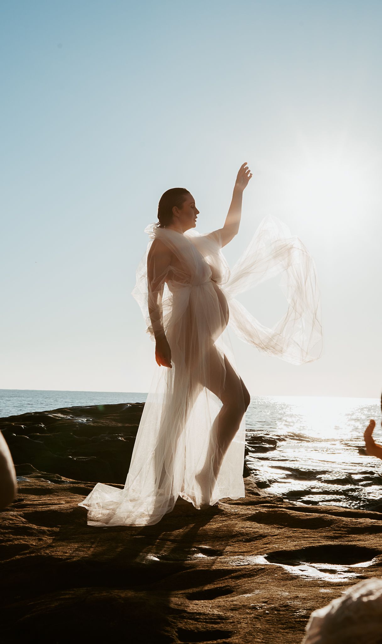 Sunlit maternity silhouette in white flowing fabric poses gracefully on rocky coastal terrain.