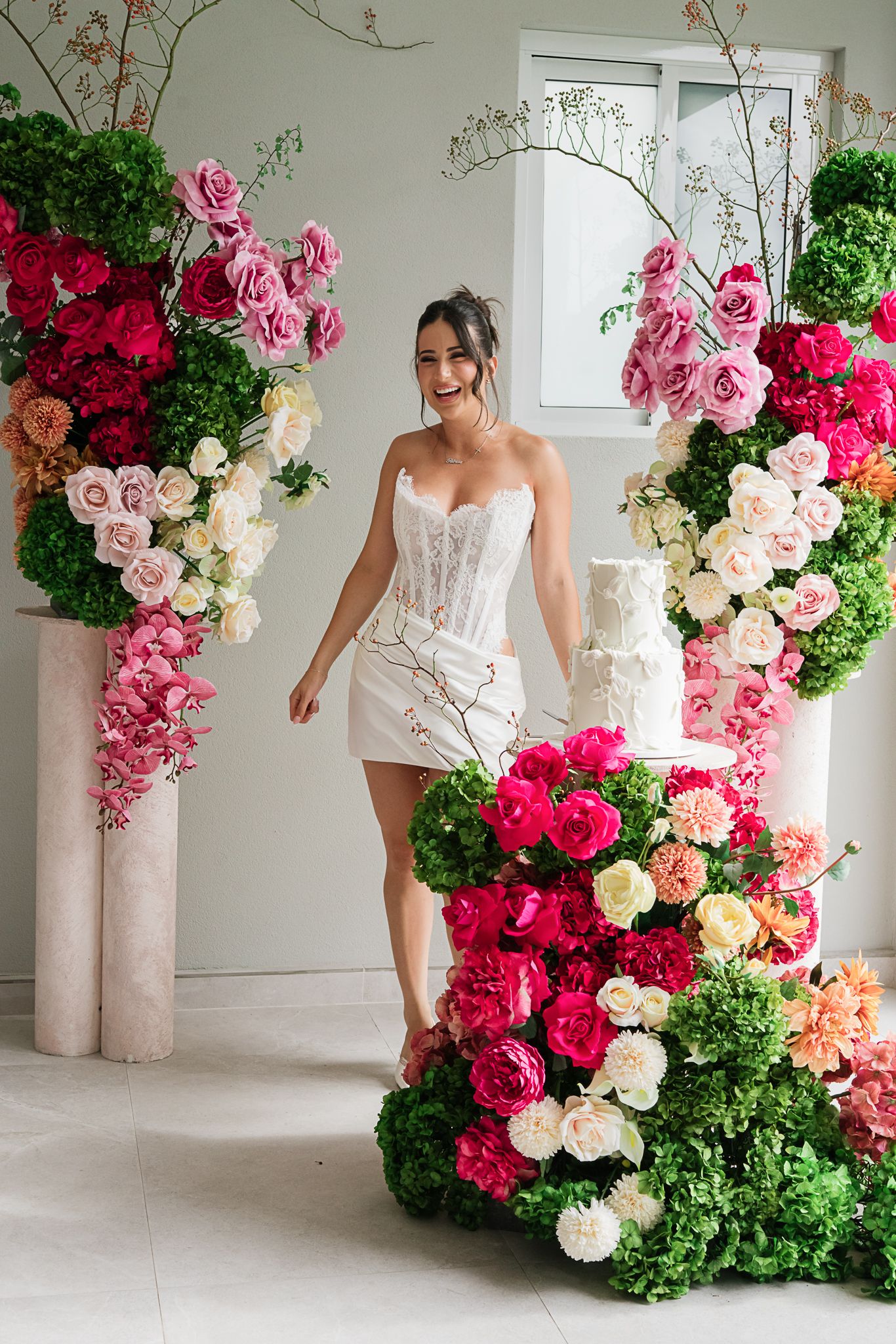 Bridal photo shoot featuring white mini dress against luxurious floral arrangements in pink and red roses with green accents.