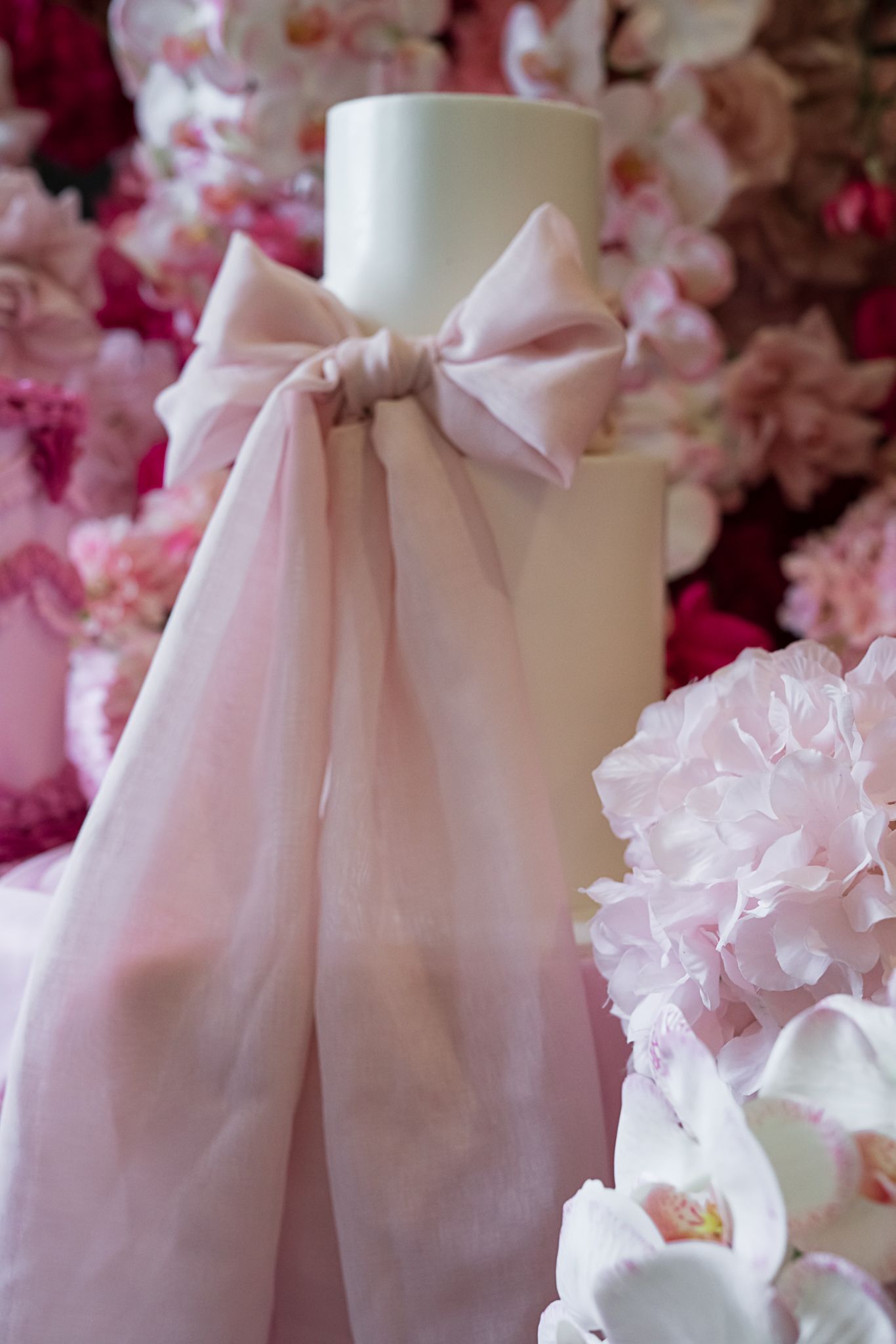 White decorative candle wrapped with soft pink tulle bow surrounded by pink floral arrangements and orchids.