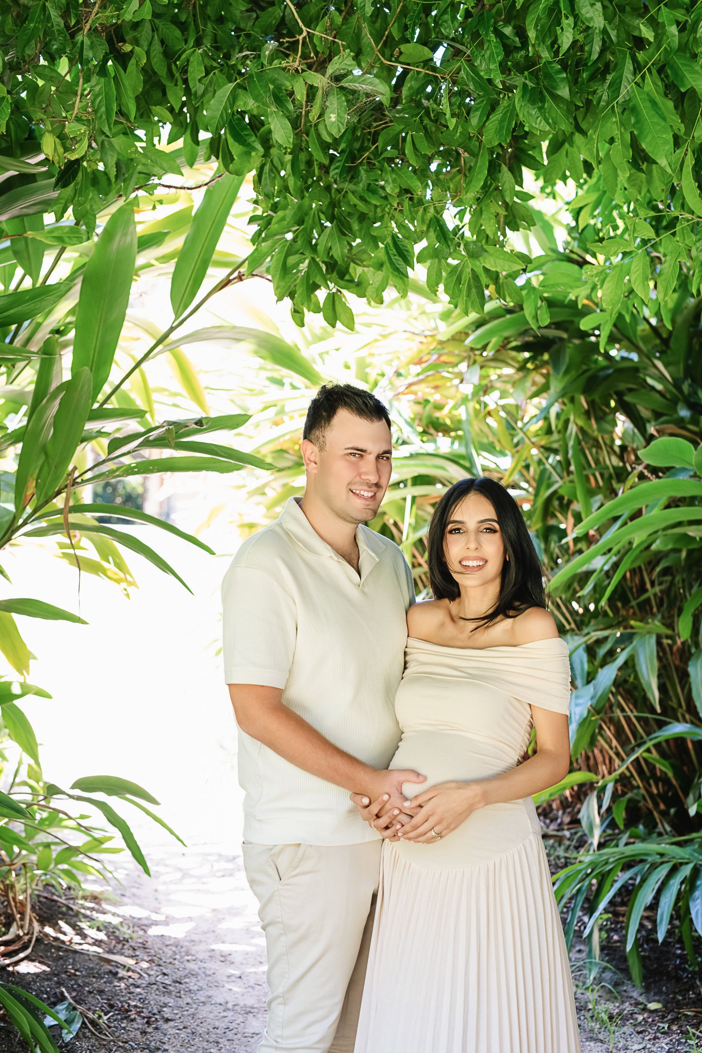 Smiling couple in light-colored outfits pose for maternity photos under green foliage.