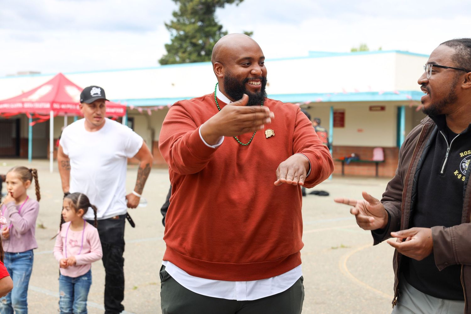 People engaged in animated conversation outdoors at a casual community gathering with red tent visible in background.