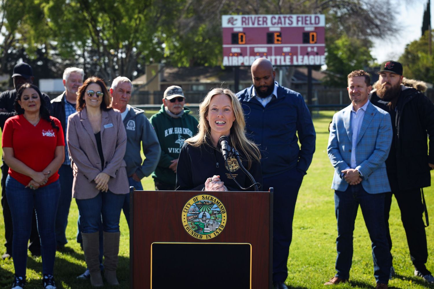 Official speaks at outdoor podium with state seal while others stand behind at public event on sunny day.