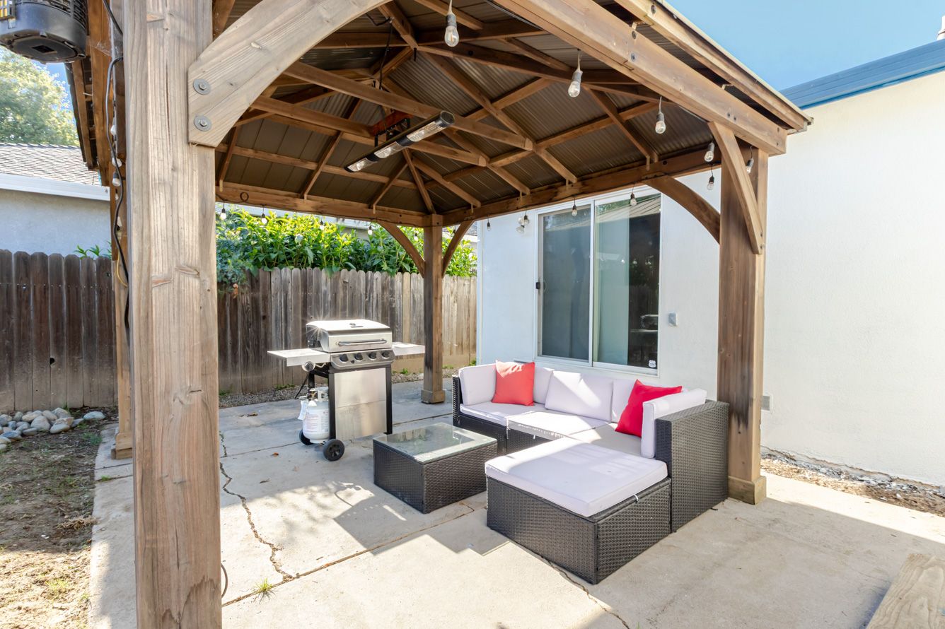 Outdoor patio with wooden gazebo, wicker sectional sofa with red pillows, and stainless steel grill on concrete pavers.