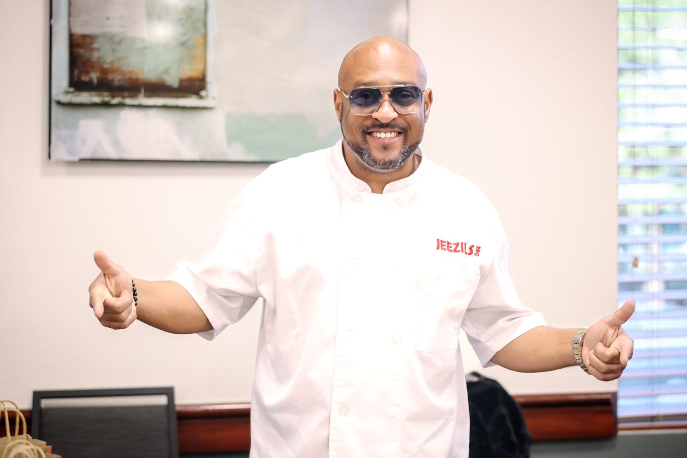 Chef in white uniform and sunglasses smiles with arms outstretched in a welcoming gesture at a restaurant.