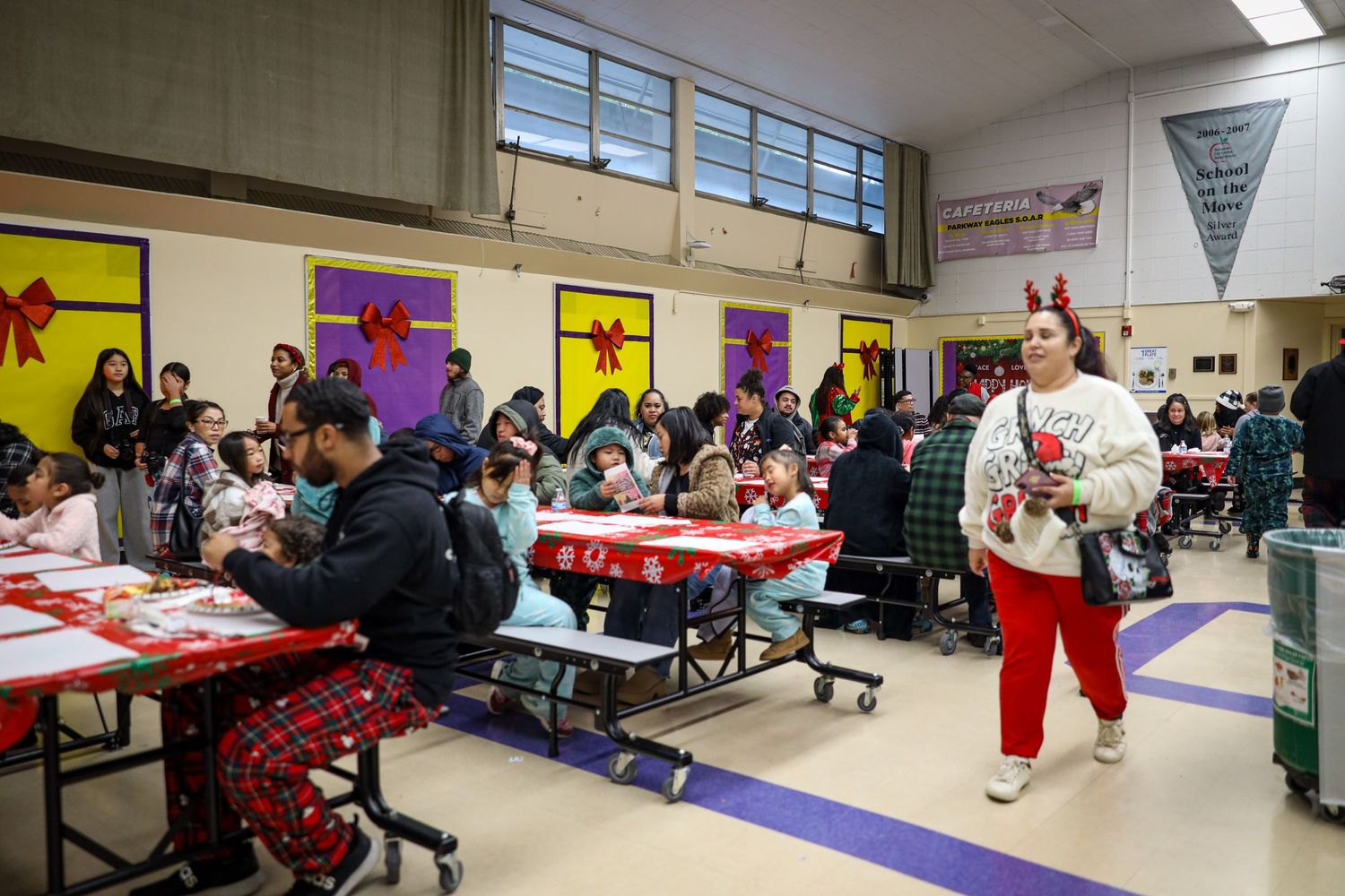 Students sit at tables in a school cafeteria during a holiday celebration with festive decorations visible.