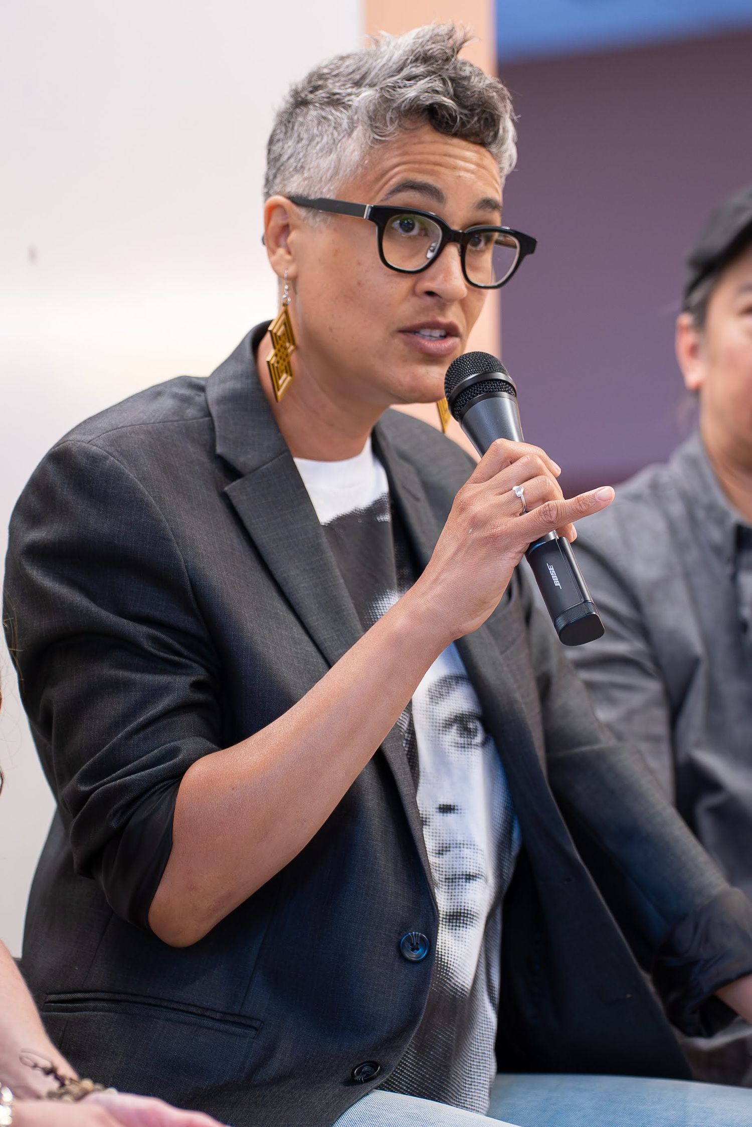 Person with gray hair and glasses speaking into a microphone at a panel event wearing a dark blazer.