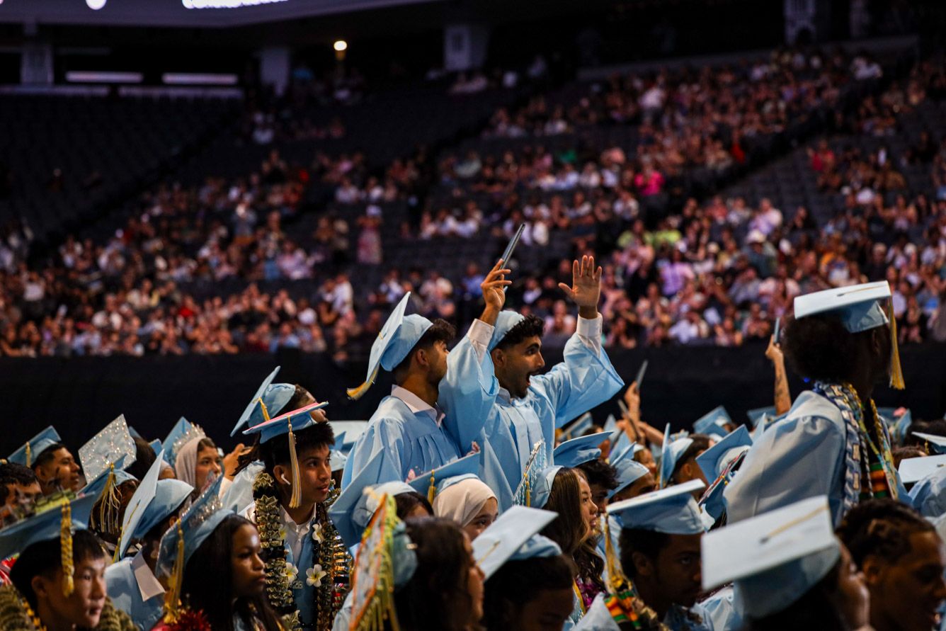 Graduates in light blue caps and gowns celebrate at a packed indoor graduation ceremony, raising hands with diplomas.