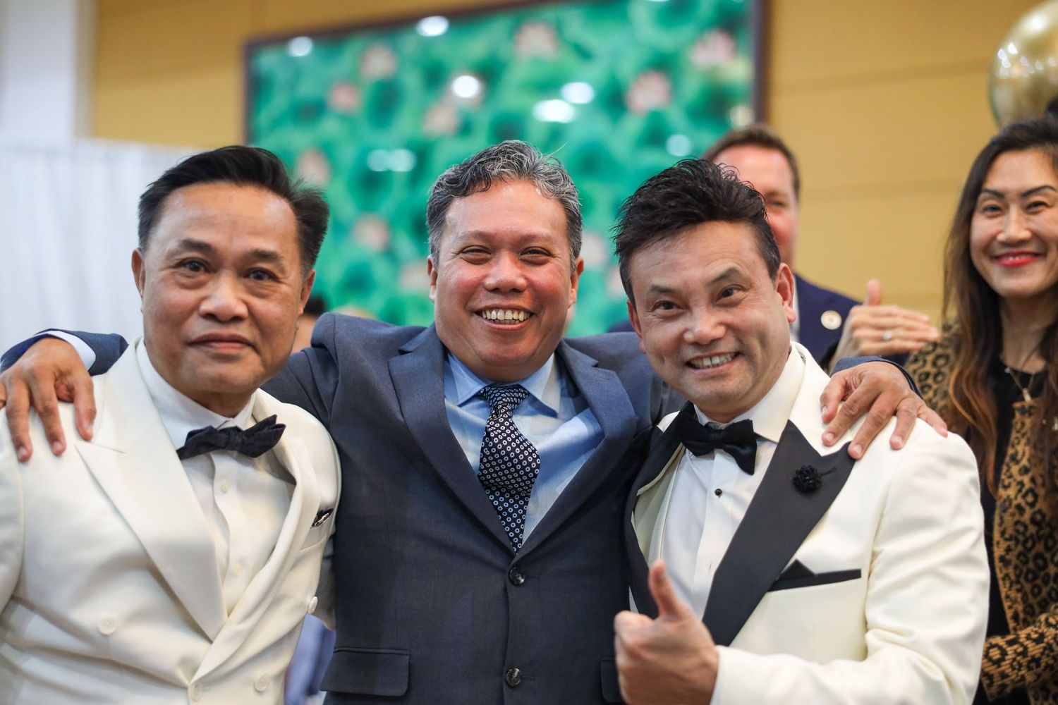 A row of formally dressed men in tuxedos and suits smile while standing together at an event.