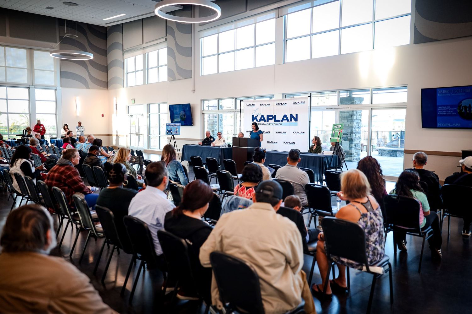 A packed conference room with people seated in rows facing a branded stage during a business presentation.