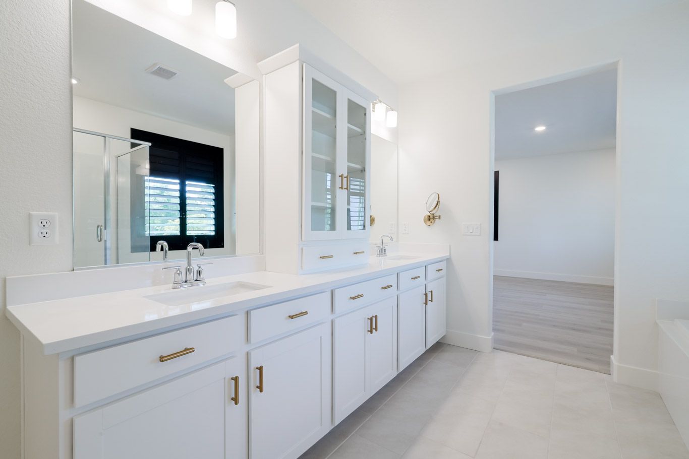Modern white bathroom with twin sinks, gold hardware, glass cabinets and quartz countertops.