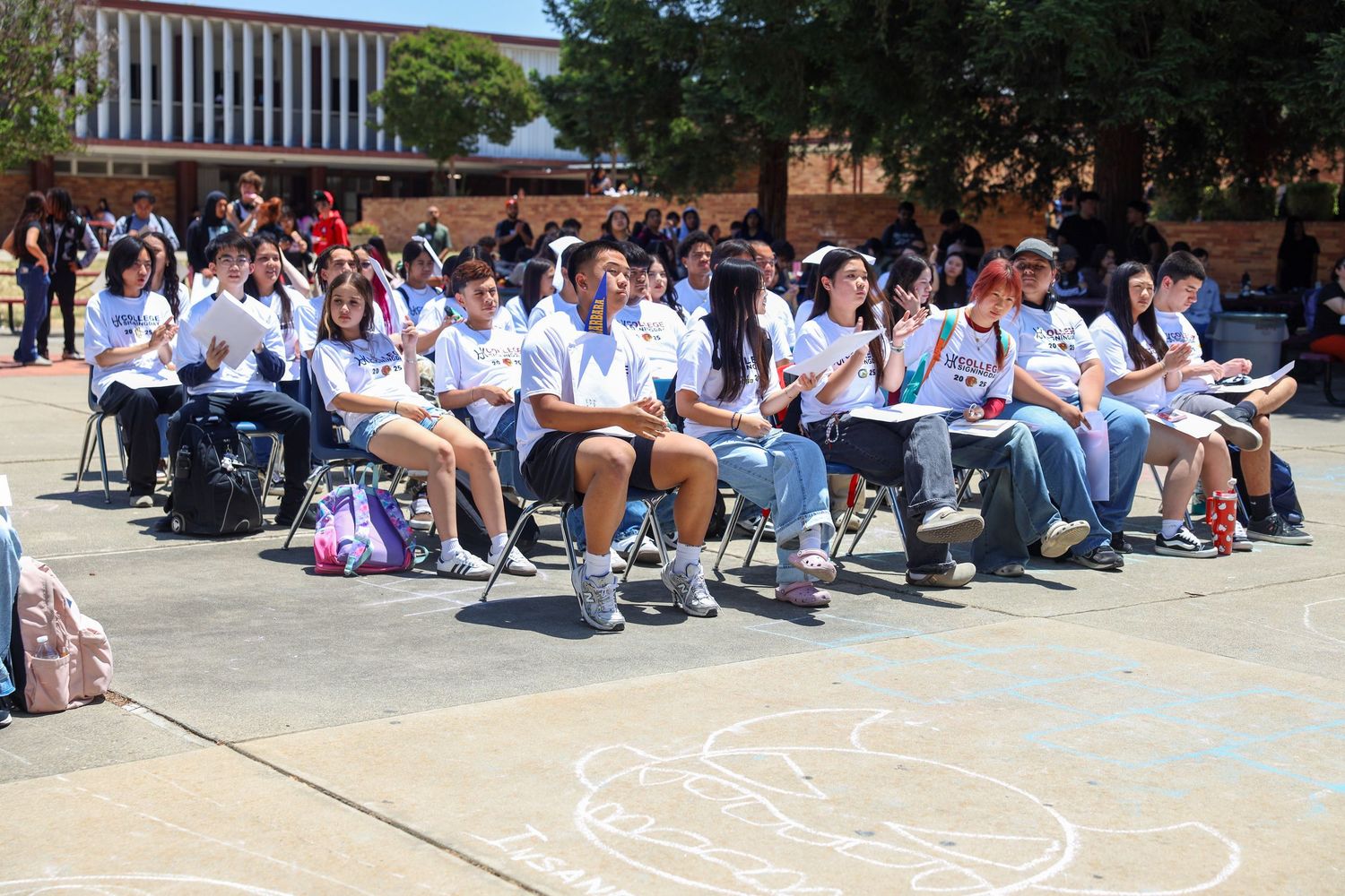 Students in blue and white uniforms sit in rows outdoors during a school event on a sunny day.