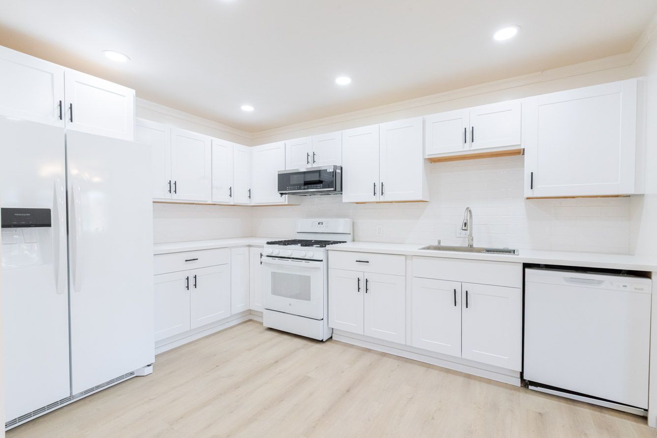 Modern white kitchen with white cabinets, white appliances, stainless steel microwave, and light wood floors.