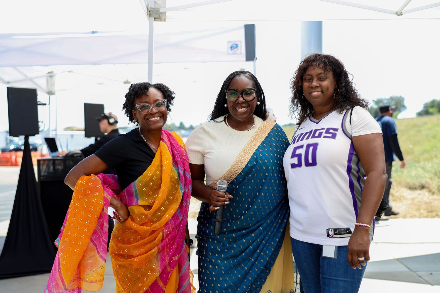 Three women smiling together outdoors at an event, wearing colorful attire including traditional Indian sarees and a Kings jersey.