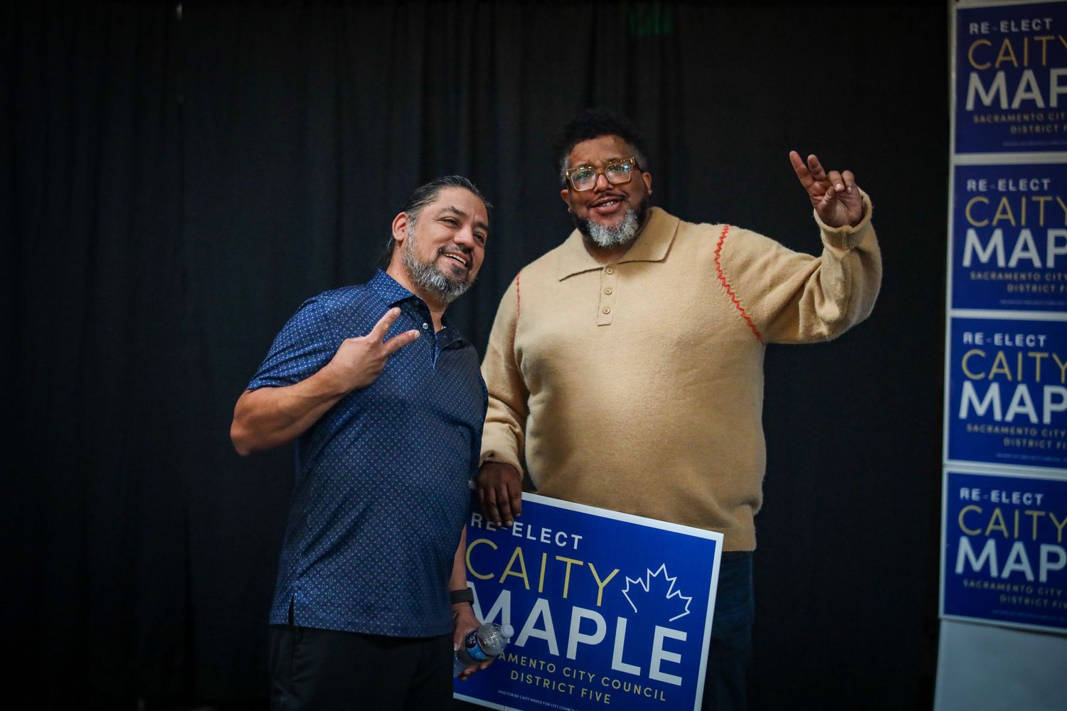 People at a political campaign event with blue and white Maple campaign signs visible in the background.