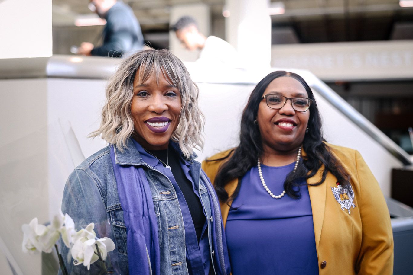 Two friends wearing denim and mustard yellow jackets smile together while standing outside with white flowers.