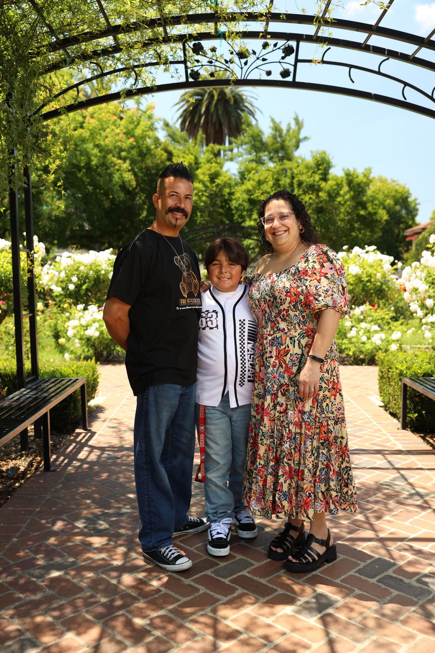 Family of three posing under decorative garden archway surrounded by lush greenery and roses on a sunny day.