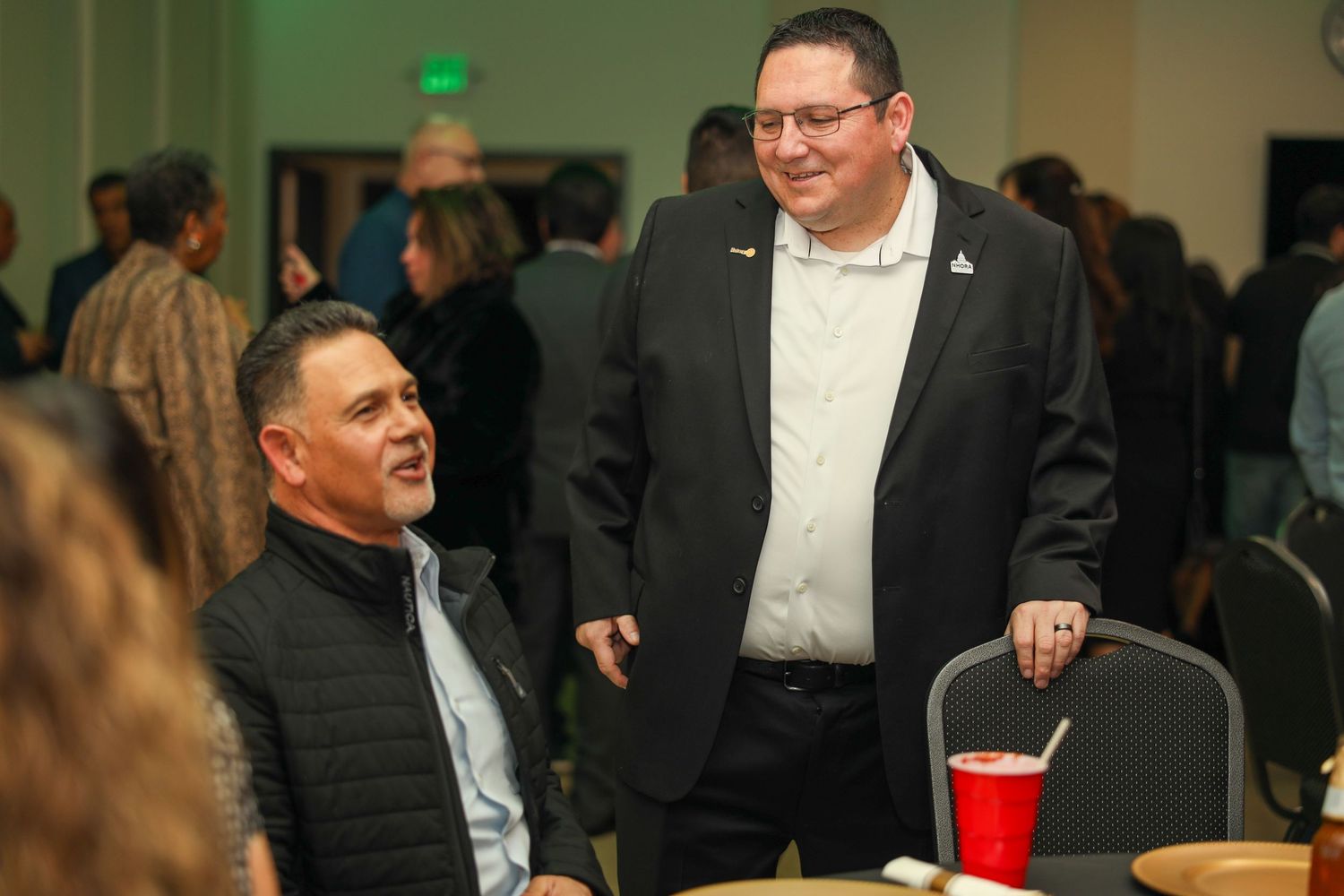 Two men in business attire having a conversation at an indoor event with people mingling in the background.