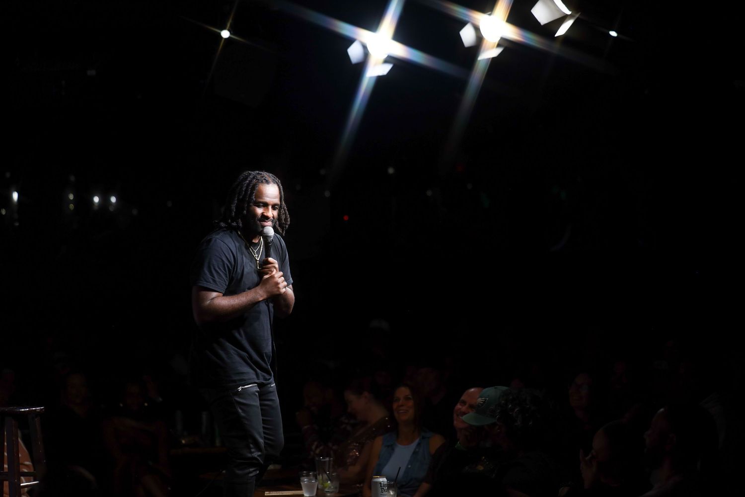 A performer stands on a dark stage with dramatic spotlights shining from above during a comedy show performance.