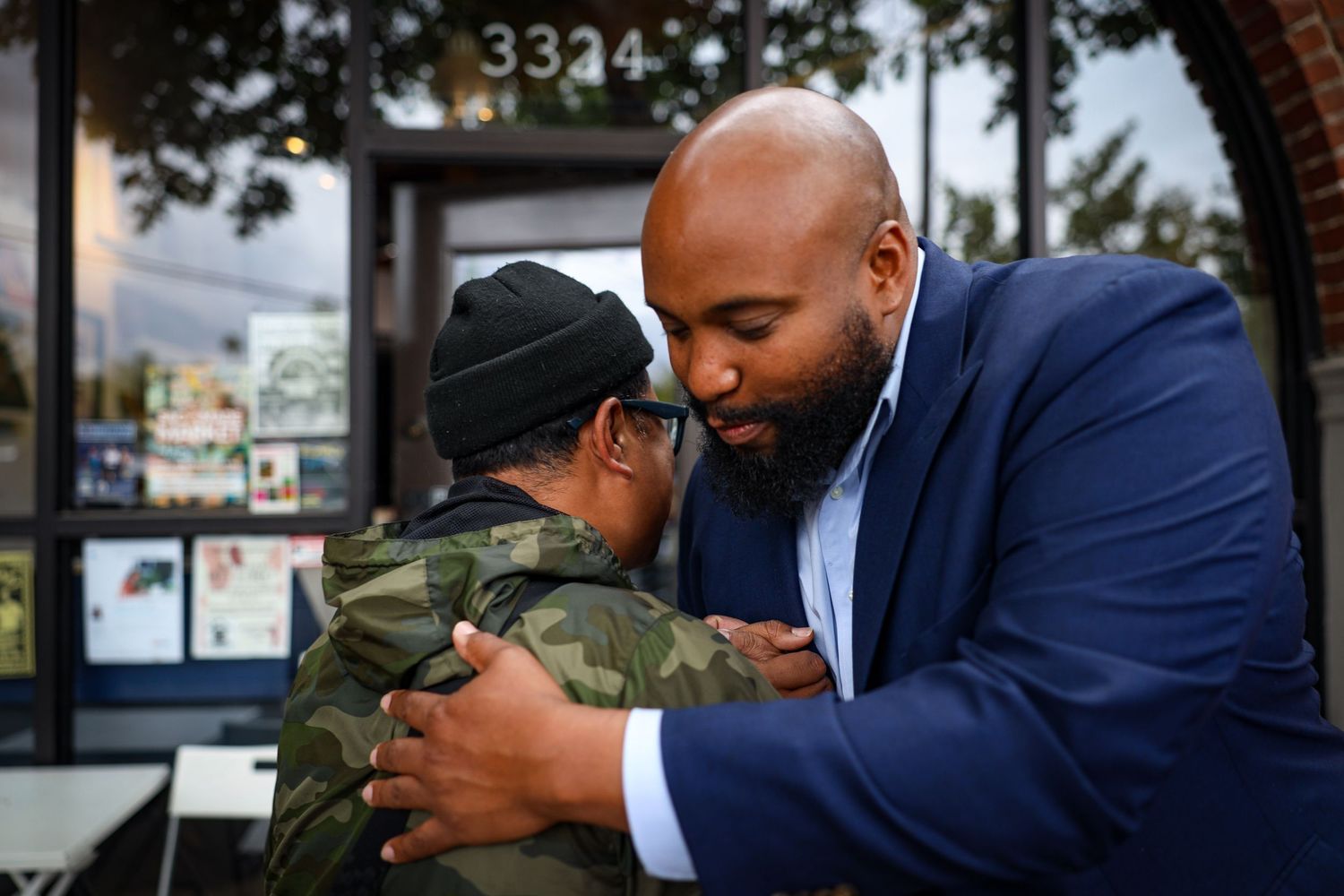 A person in a navy suit embraces someone wearing camouflage and a beanie outside a storefront.