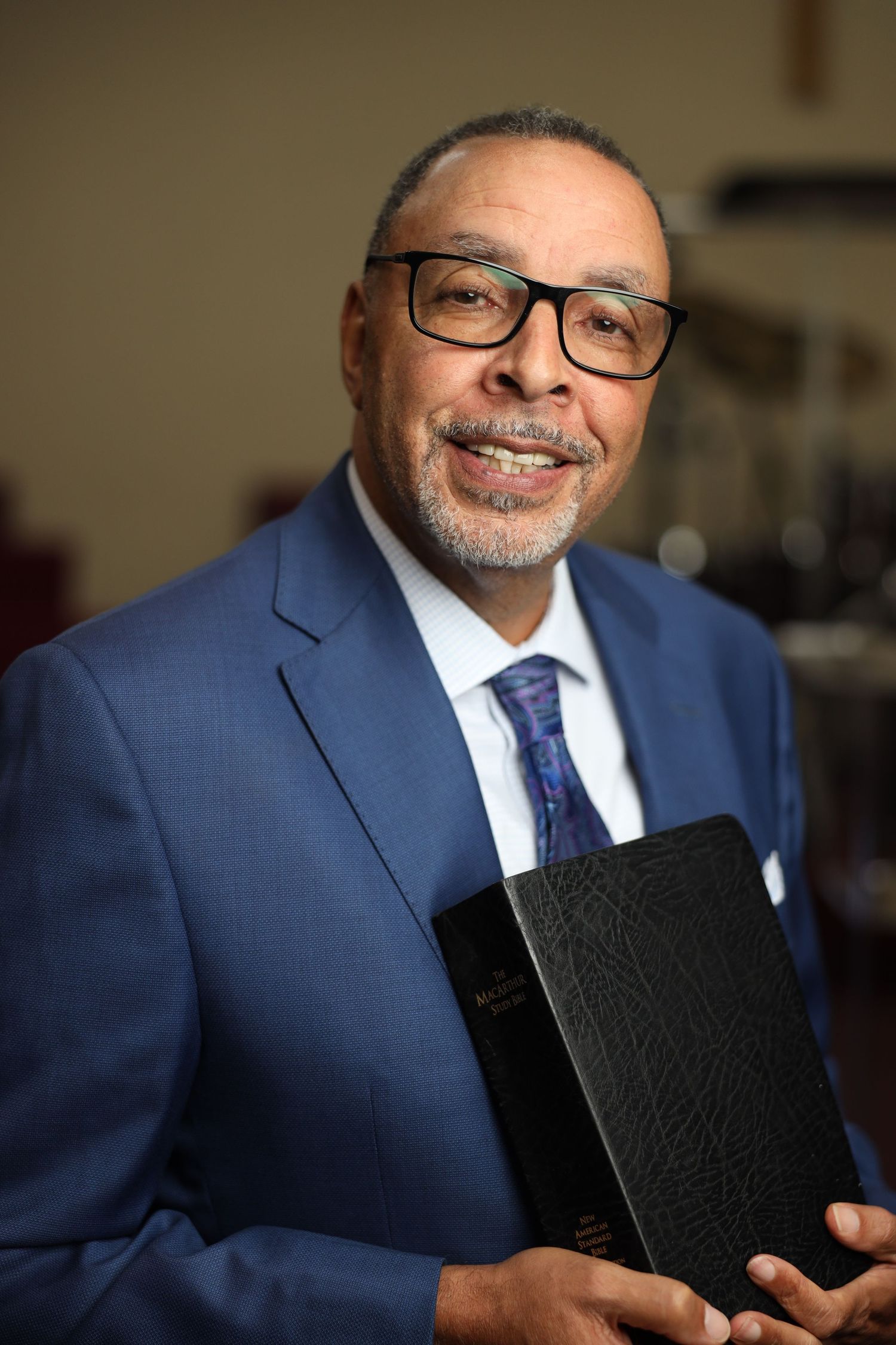Professional portrait of someone in a navy blue suit and glasses holding a black leather portfolio with a warm smile.