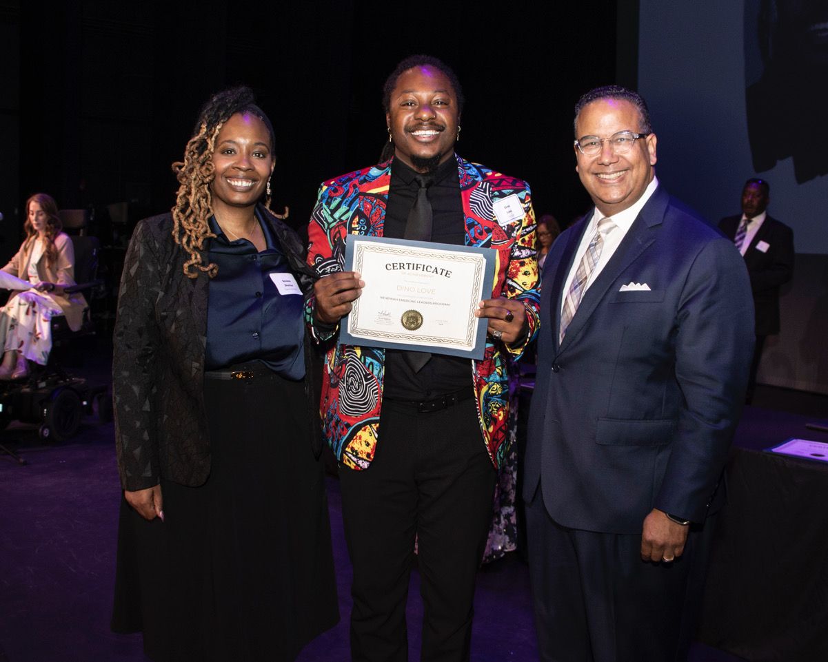 Three people pose together at a formal awards ceremony, with the center person holding a certificate of achievement.