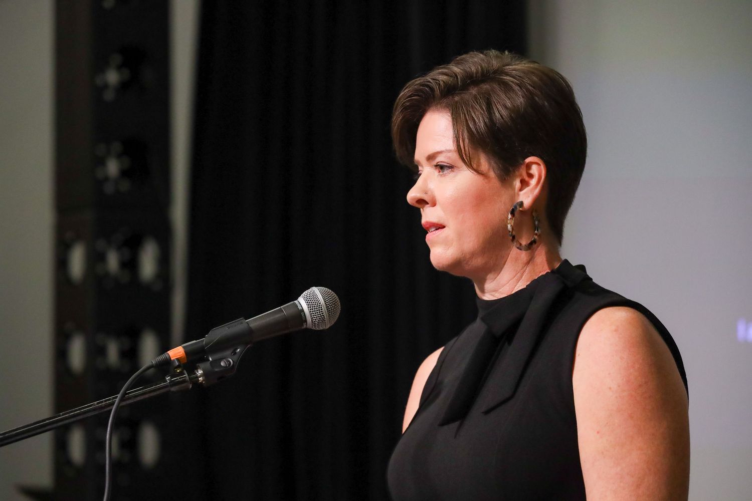 Woman in black dress speaking at a microphone podium during a formal event or conference.