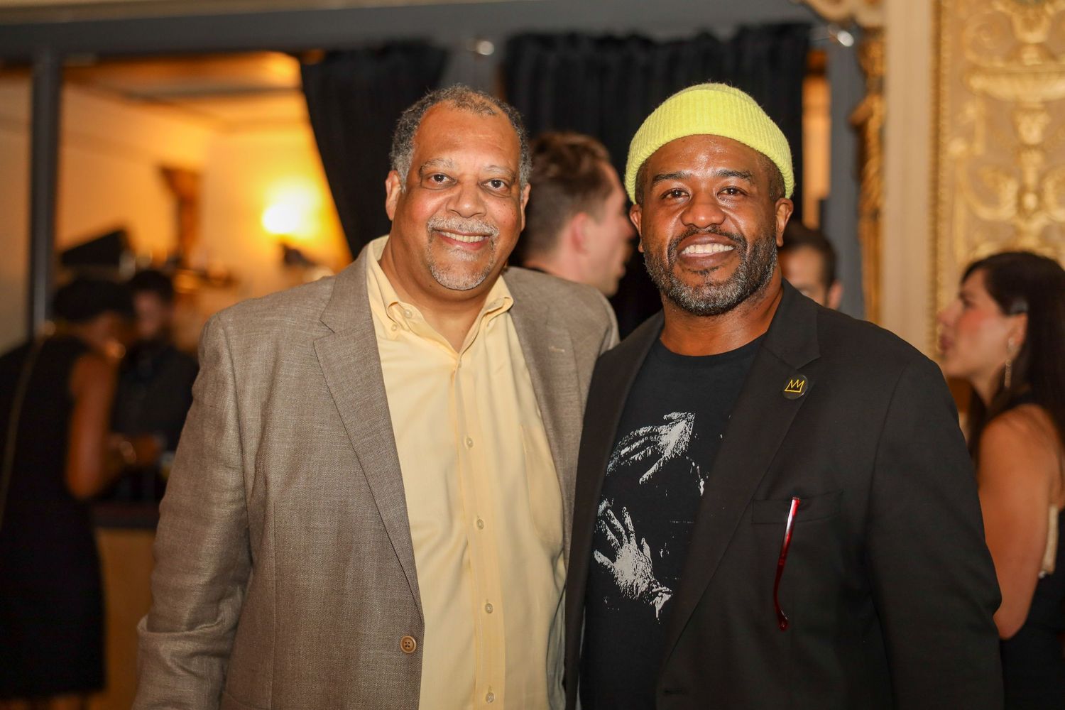 Two smiling men pose together at an indoor event, one in a tan blazer and one in a black outfit with a green hat.