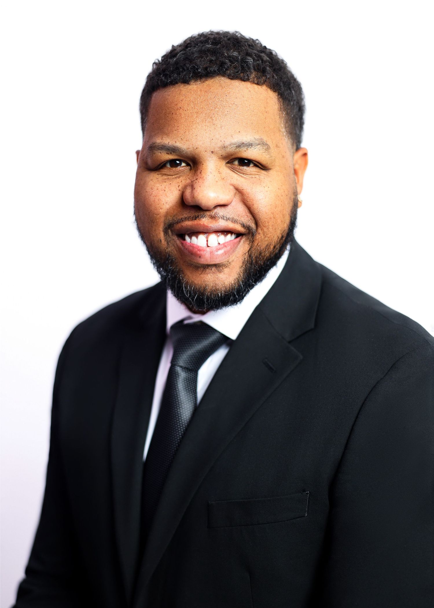 A person in a black suit and tie smiles warmly against a light background.
