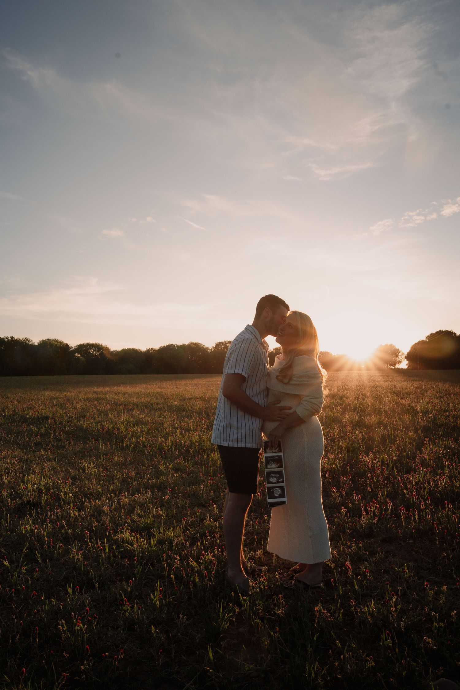 Couple embracing in golden sunset field holding ultrasound photos, announcing pregnancy outdoors.
