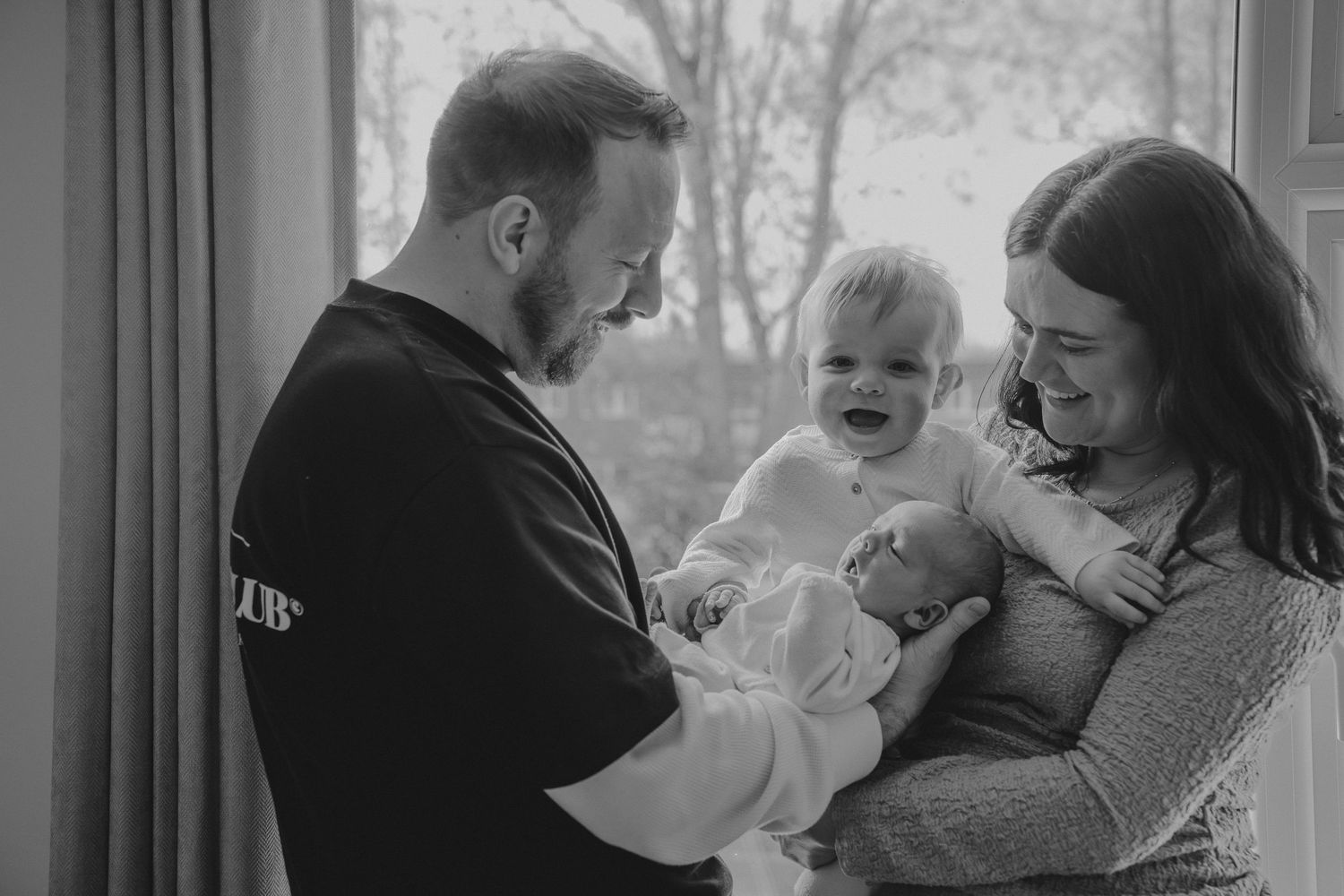 Black and white photo of a couple smiling at their newborn baby while a toddler laughs joyfully near a window.