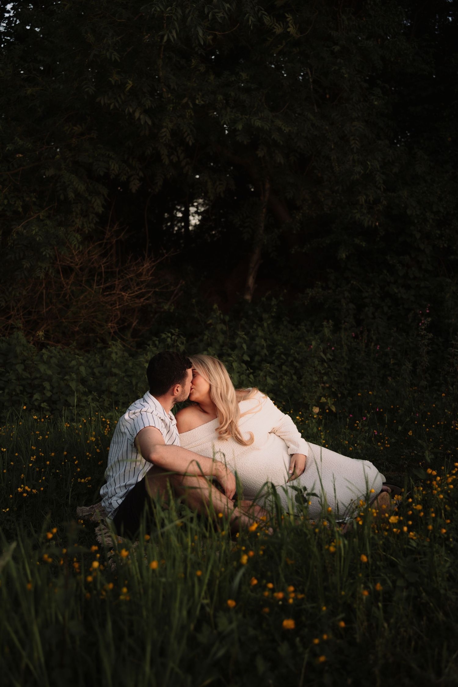 Couple sharing a kiss while sitting in a wildflower meadow surrounded by lush green trees at golden hour.
