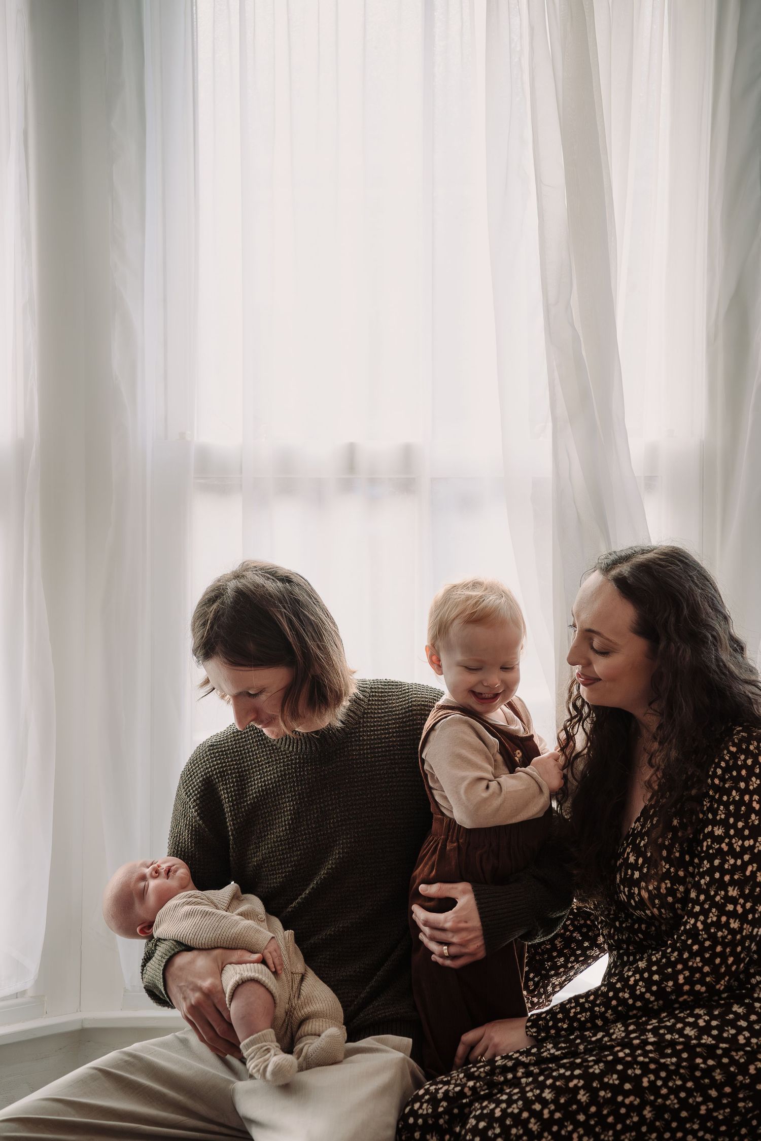 An intimate family portrait series showing loving interactions between parent and infant by a window.
